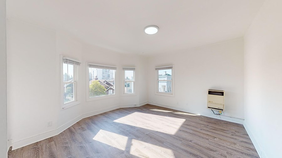 A room with wooden floors and white walls, with a window on the left and right, and a small air conditioning unit on the floor.