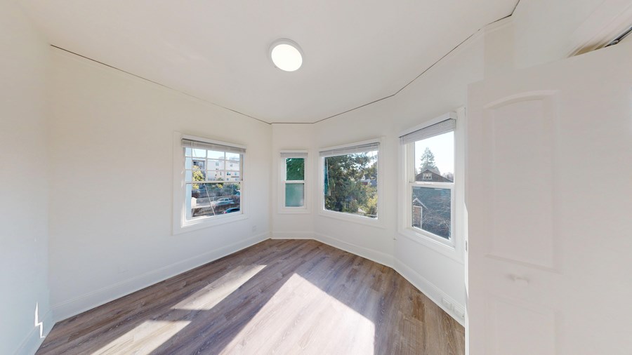 A room with white walls and wooden flooring, with three windows showing an outdoor view.