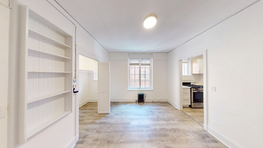 A spacious, empty room with a window and a door leading to a kitchen.