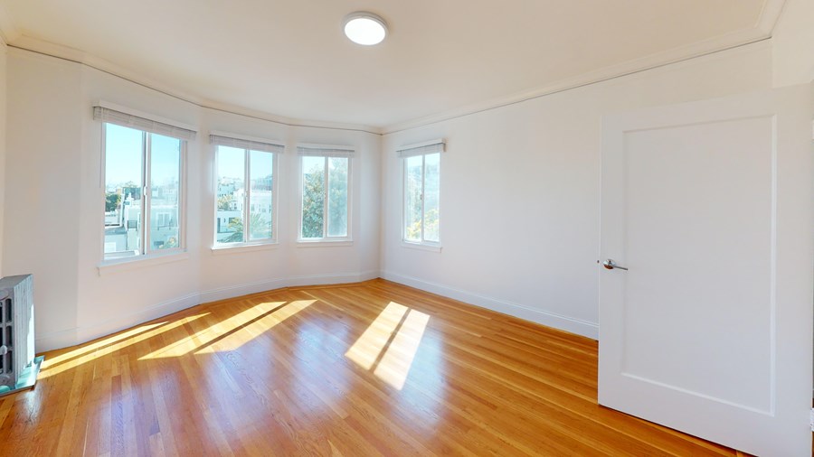 A room with wooden floors and white walls has a white door and a window with a view of trees.