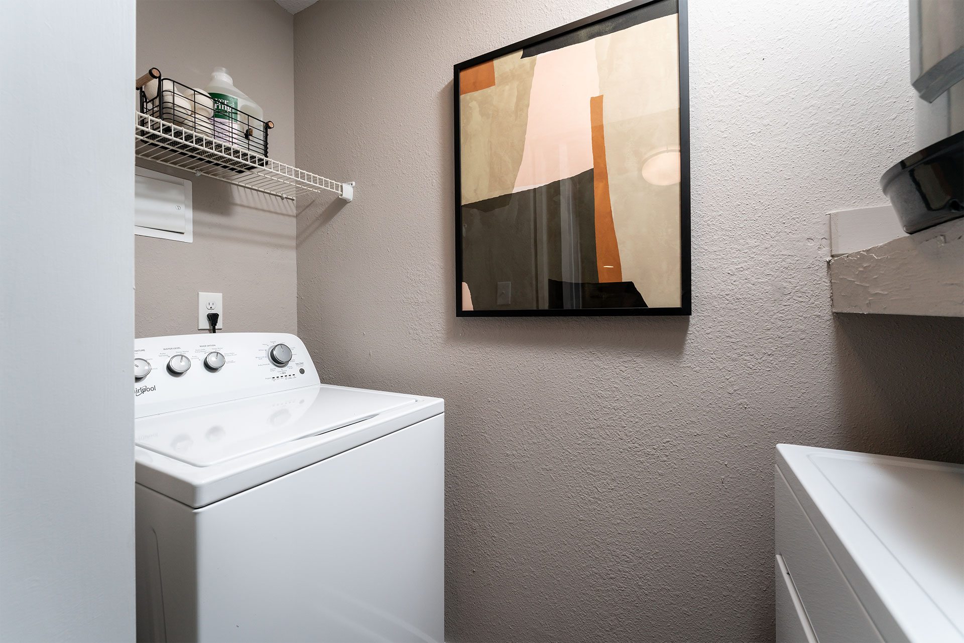 A washing machine sits in a laundry room with a painting hanging on the wall.