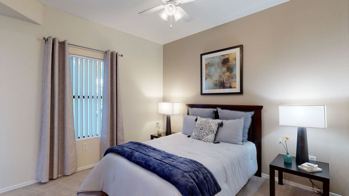 A cozy and tranquil bedroom here at Pala Mesa featuring a plush bed with layered neutral bedding and a soft accent throw, framed by matching bedside tables with modern lamps. A large window with vertical blinds and curtains allows natural light to filter in, while plush carpeting and calming wall tones create a peaceful, comfortable retreat.