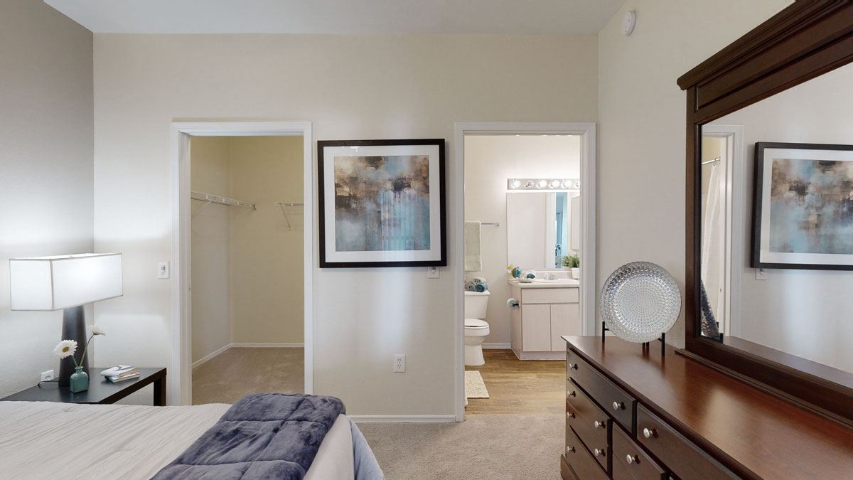 A spacious and inviting bedroom here at Pala Mesa featuring soft neutral walls, plush carpeting, and a comfortable bed with layered bedding and accent throw. The room opens to a generous walk-in closet and a private bathroom with a large vanity and bright mirror lighting, while a coordinating dresser and bedside table add warmth and functional storage.