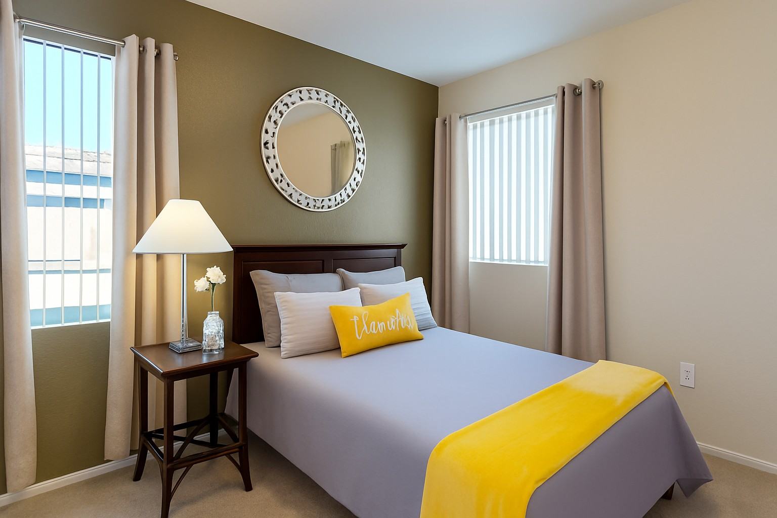 A warm and inviting bedroom here at Pala Mesa featuring soft carpeting, a rich wood headboard, and neutral bedding accented with a vibrant yellow throw and decorative pillow. A bedside table with lamp adds cozy lighting, while two tall windows with vertical blinds and curtains fill the space with natural light.