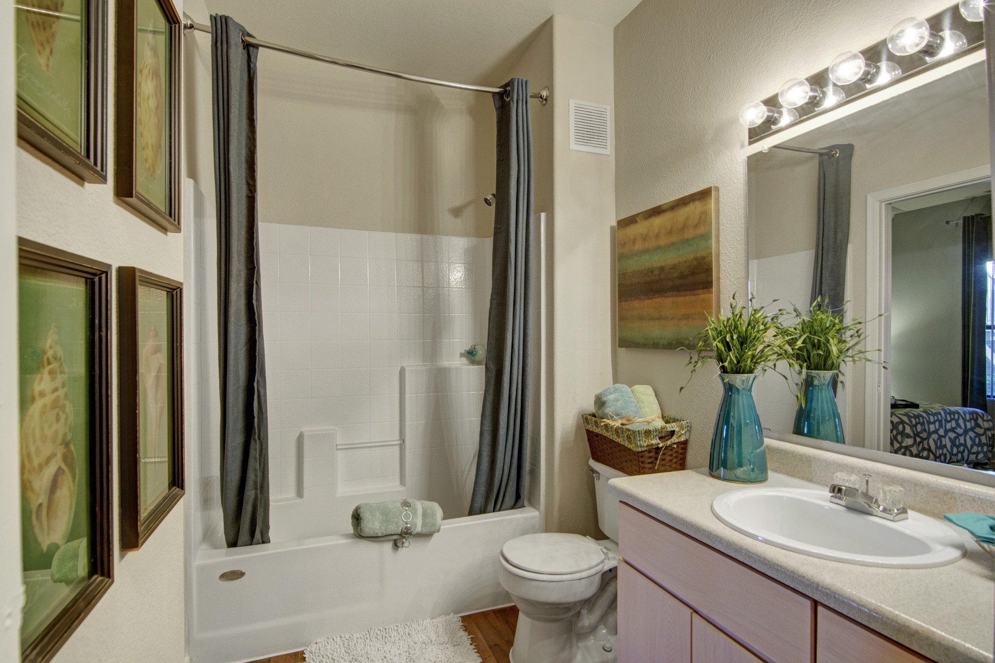 A bright and well-appointed bathroom here at Pala Mesa featuring a full tub and shower combination with tile surround and a curved curtain rod. A spacious vanity with ample counter space and a large mirror is complemented by modern lighting, while decorative accents and soft towels create a clean and inviting atmosphere.