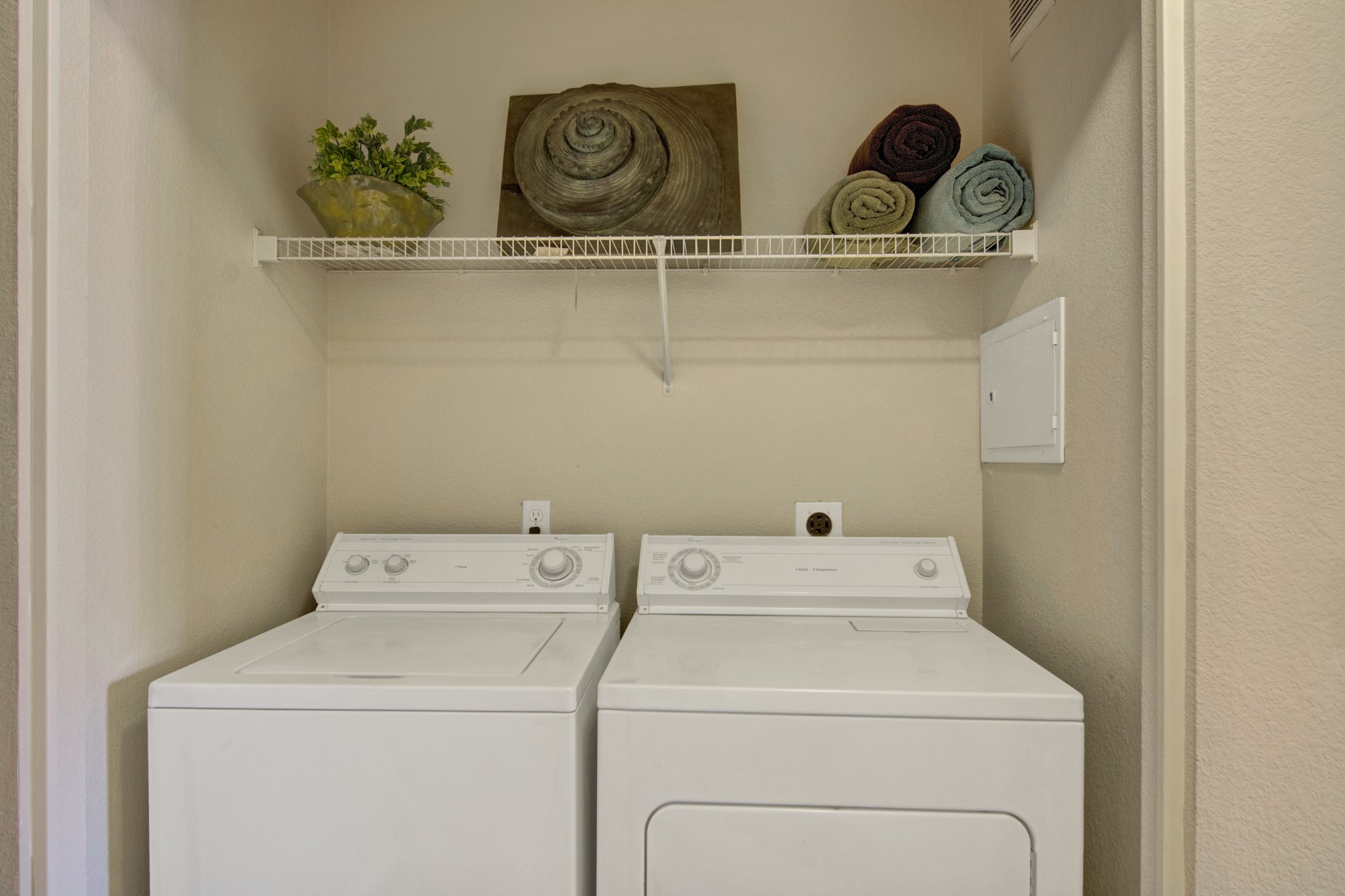 A convenient in-home laundry area here at Pala Mesa featuring a full-size washer and dryer set neatly tucked into a dedicated closet space. A wide wire shelf above provides additional storage for linens and essentials, while neutral walls create a clean, organized setting designed for everyday comfort and functionality.