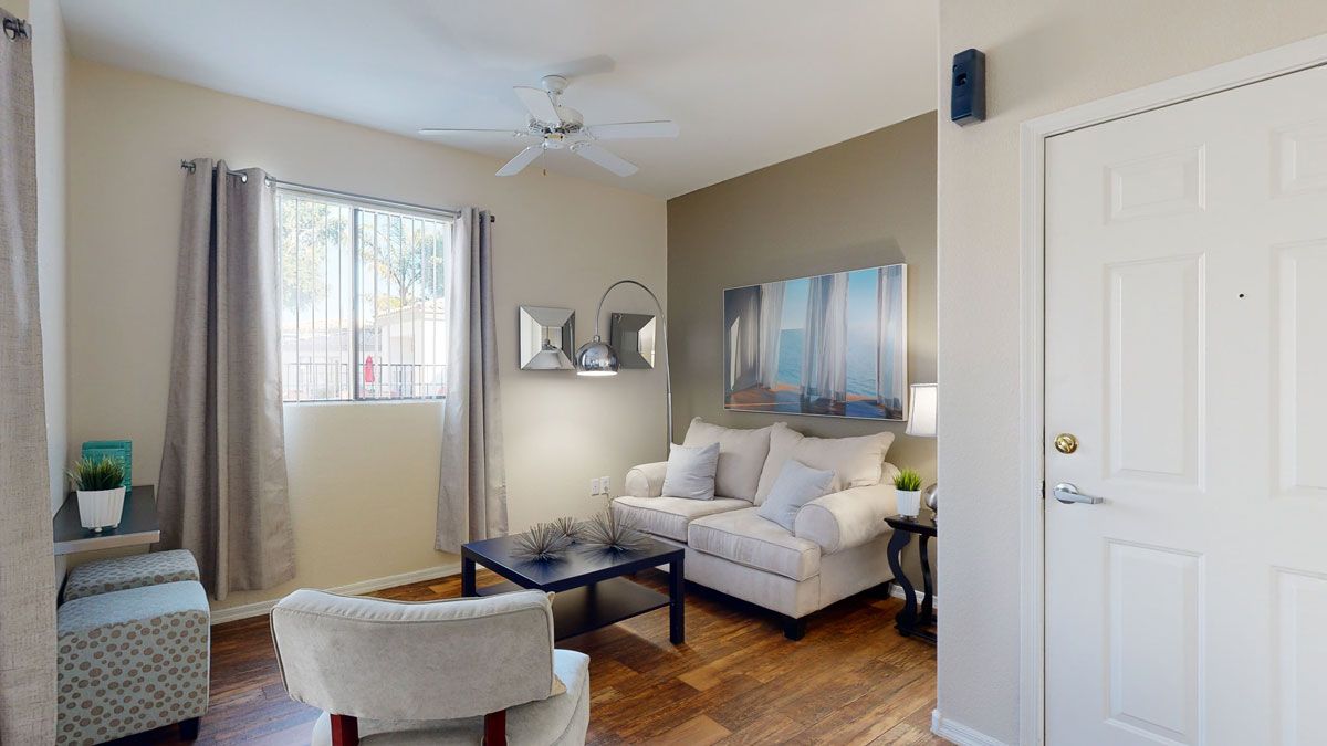 A bright and inviting living room here at Pala Mesa featuring warm wood-style flooring, a plush cream sofa with soft accent pillows, and a sleek dark coffee table centered between cozy seating. A large window with curtains brings in natural light, while modern wall art and a stylish floor lamp create a comfortable and welcoming atmosphere near the entryway.