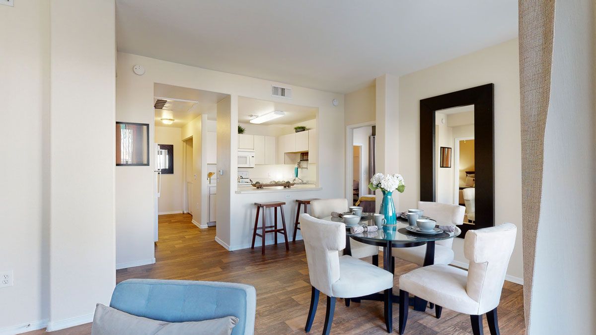 A bright and welcoming dining area here at Pala Mesa featuring rich wood-style flooring, a round glass-top table set for four with upholstered cream chairs, and a bold turquoise vase centerpiece. The open pass-through to the kitchen includes a breakfast bar with stools, while neutral walls and a large framed mirror create an airy, spacious feel.
