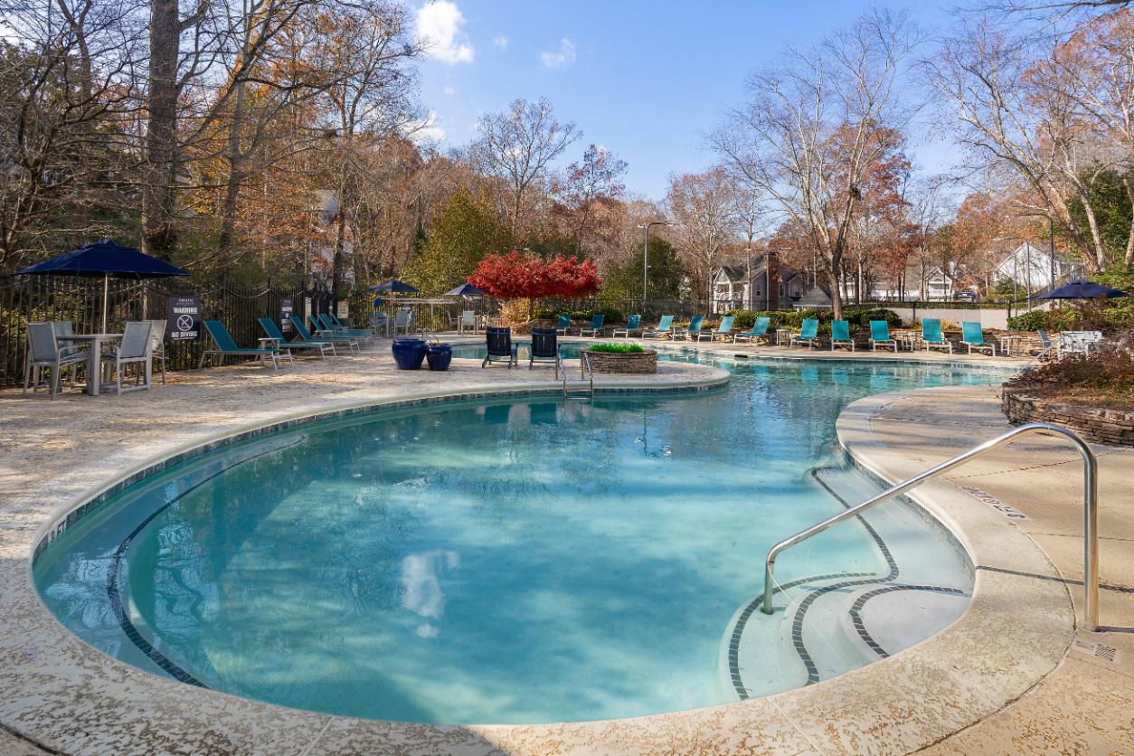 A large outdoor swimming pool surrounded by trees and a patio area with chairs and umbrellas.