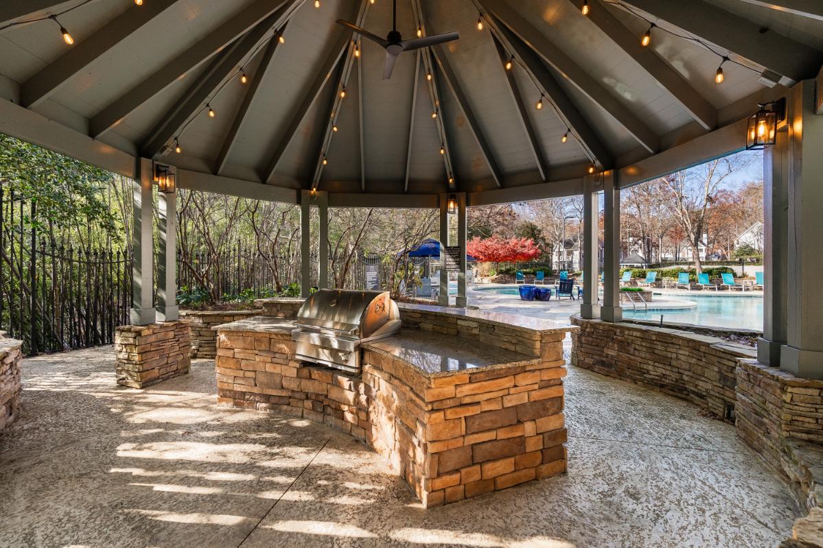 A gazebo with a stone bench and a fireplace is surrounded by a gravel area.