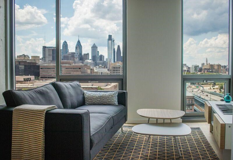 A black couch with a white pillow and a white coffee table in front of a window with a city view.