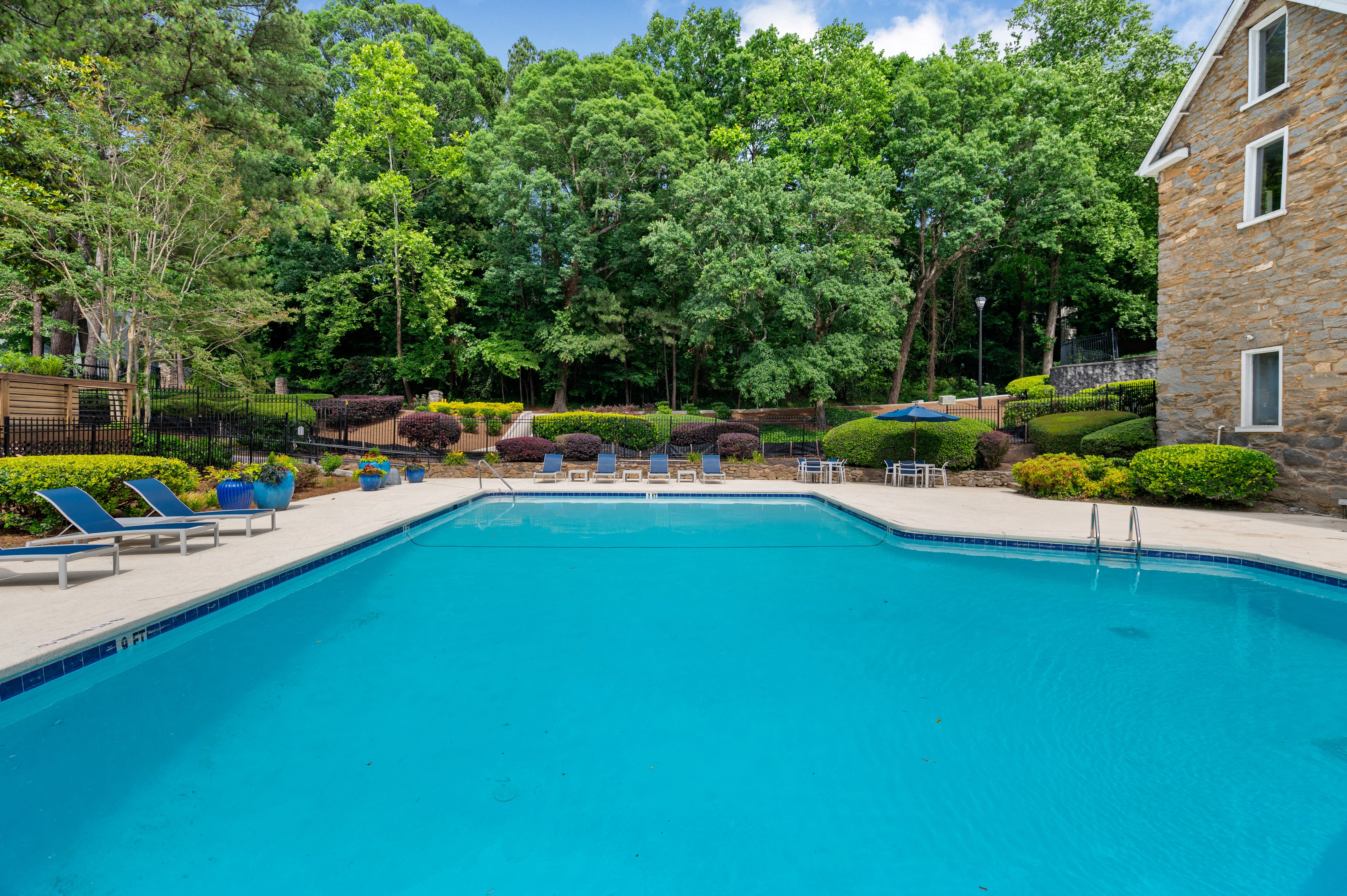 A blue swimming pool with lounge chairs next to a stone building and lush trees at Mill House at East Cobb offers townhomes in Marietta, GA.