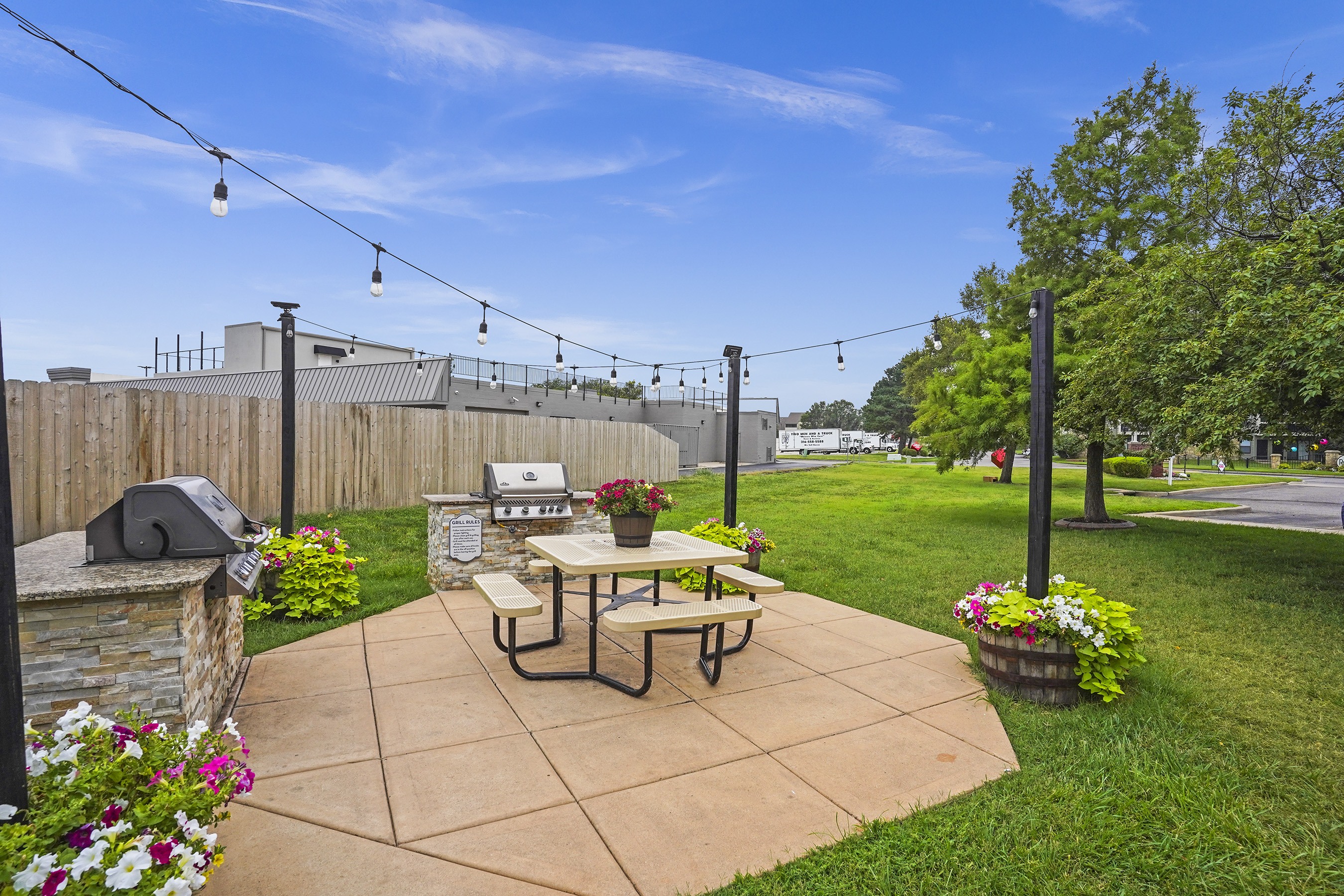 A patio with a picnic table and chairs is surrounded by a fence and has string lights above it.