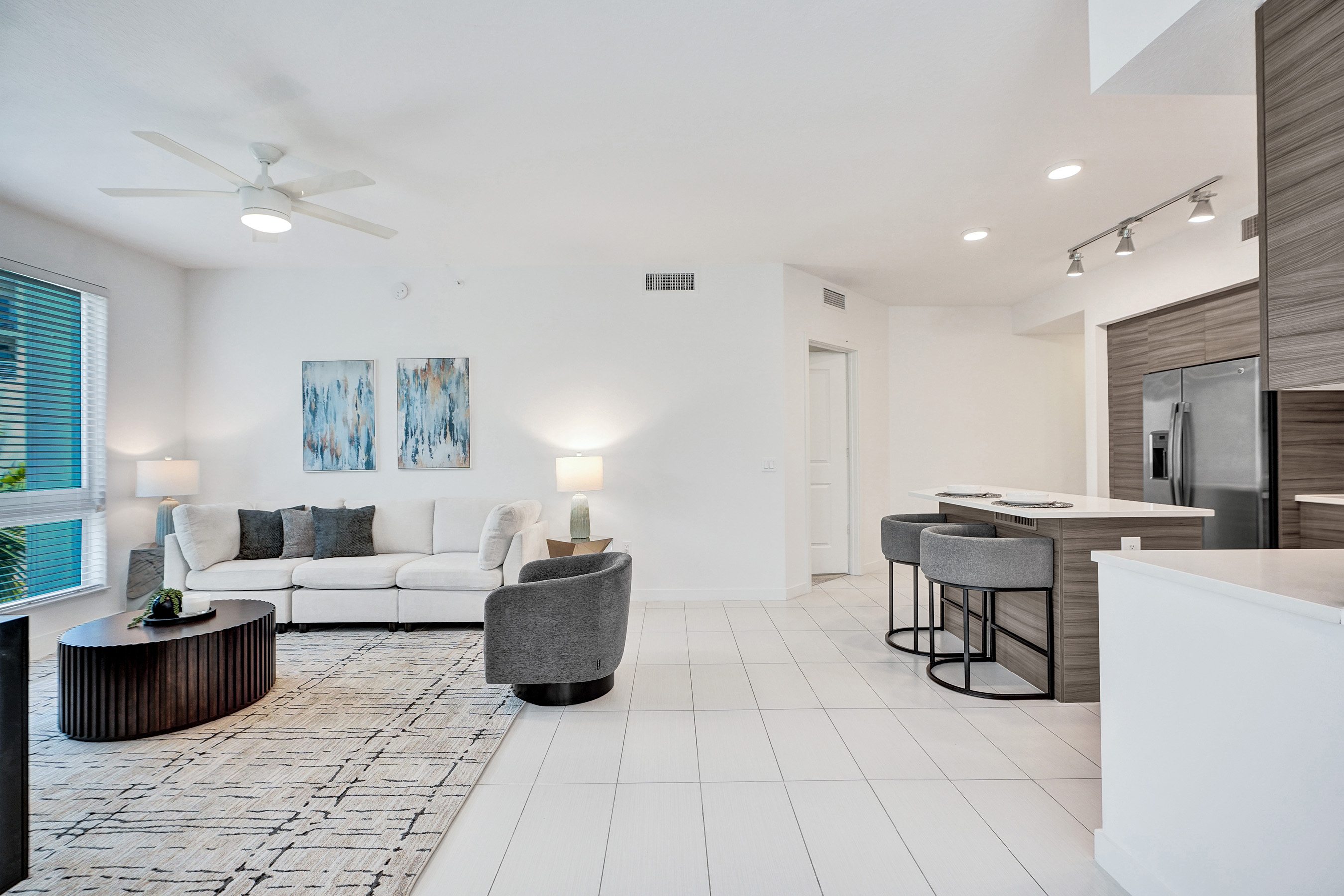 A modern living room with a white sofa, a grey armchair, a wooden coffee table, and a white kitchen area.