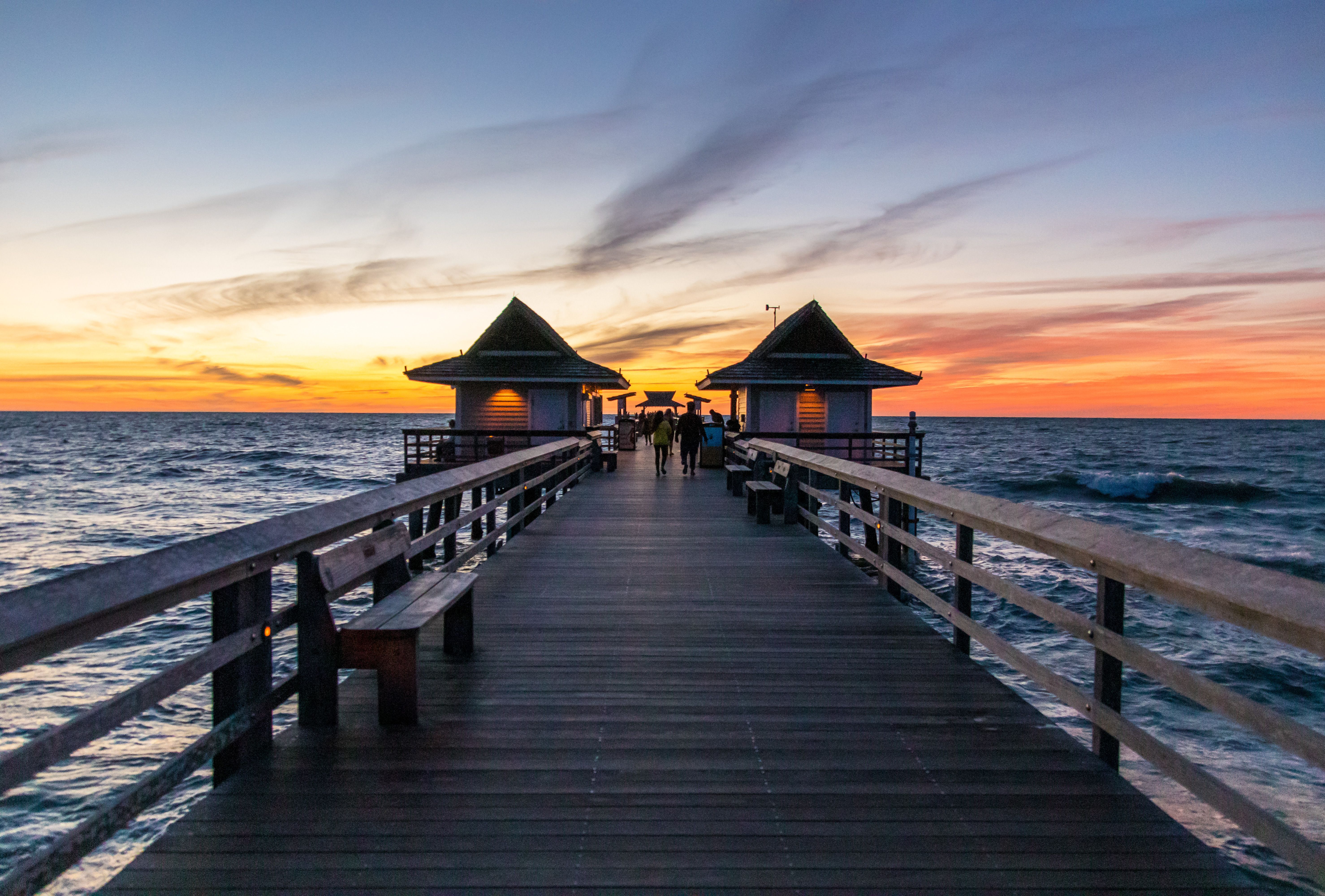 A wooden pier with two pavilions at either end extends into the ocean at sunset.